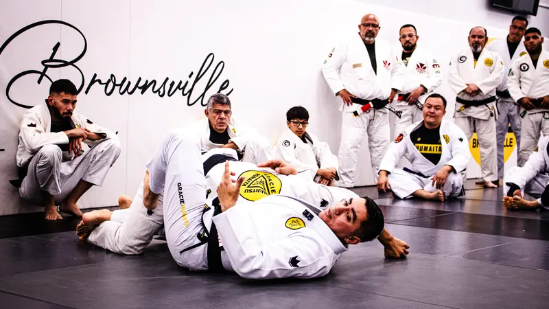 Adult students in white gis watch from the edge of the mat while an instructor demonstrates a ground technique, under the word Brownsville painted on the wall