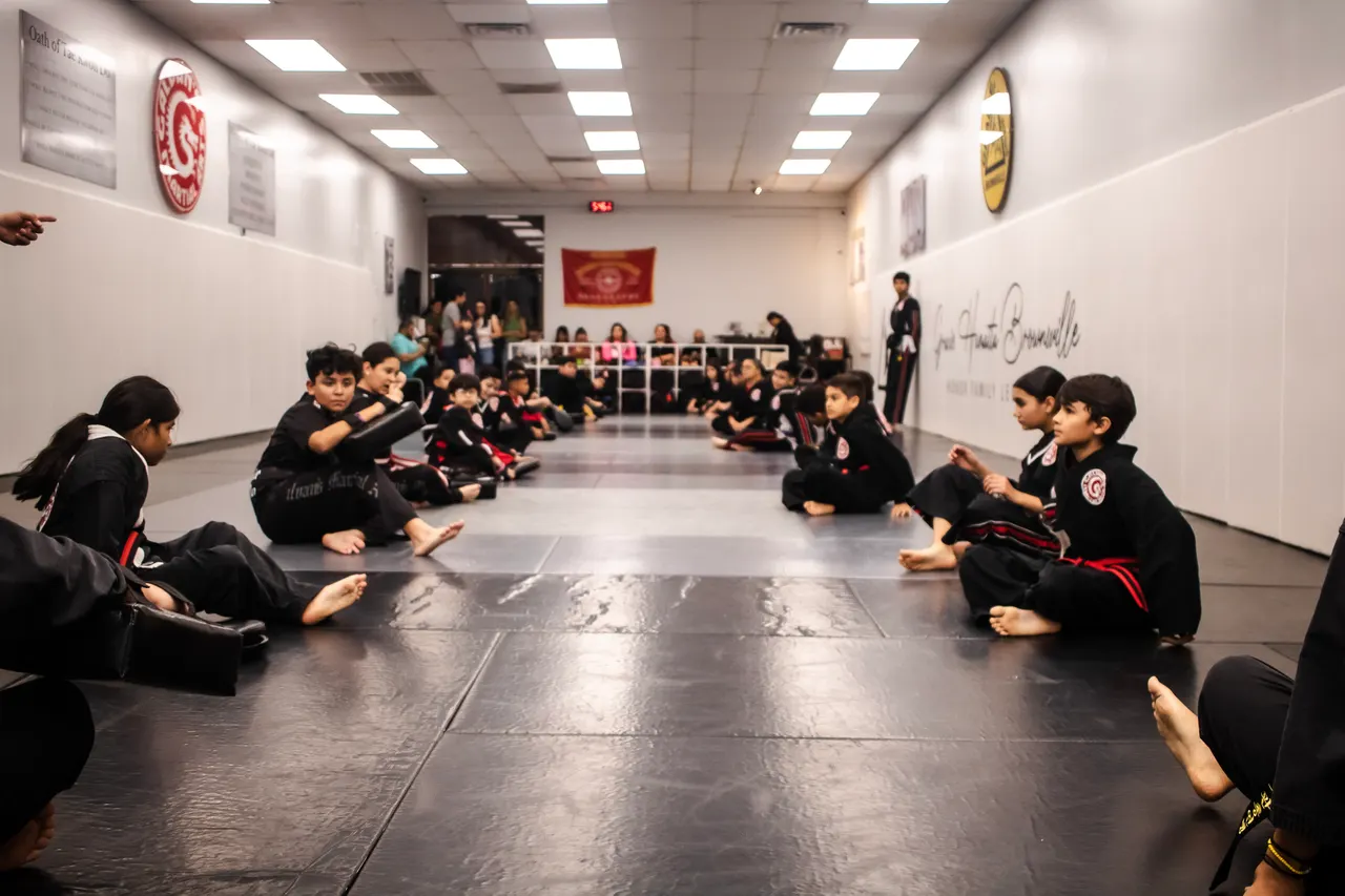 Niños con uniforme de Galvan's Martial Arts sentados a los lados del tapete durante una clase de taekwondo, con los papás observando desde el fondo