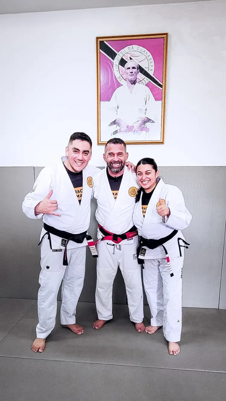 Professor Manny Galvan and Professor Martha Galvan, both in white gis with black belts, standing either side of Professor Paulo Brandao wearing his red-and-black coral belt at the Gracie Humaita headquarters in Rio de Janeiro