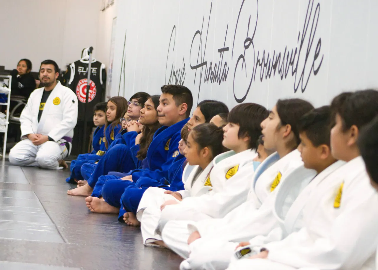 Children in blue and white jiu-jitsu gis sitting in a row along the wall listening to their instructor during a kids class at Galvan’s Martial Arts