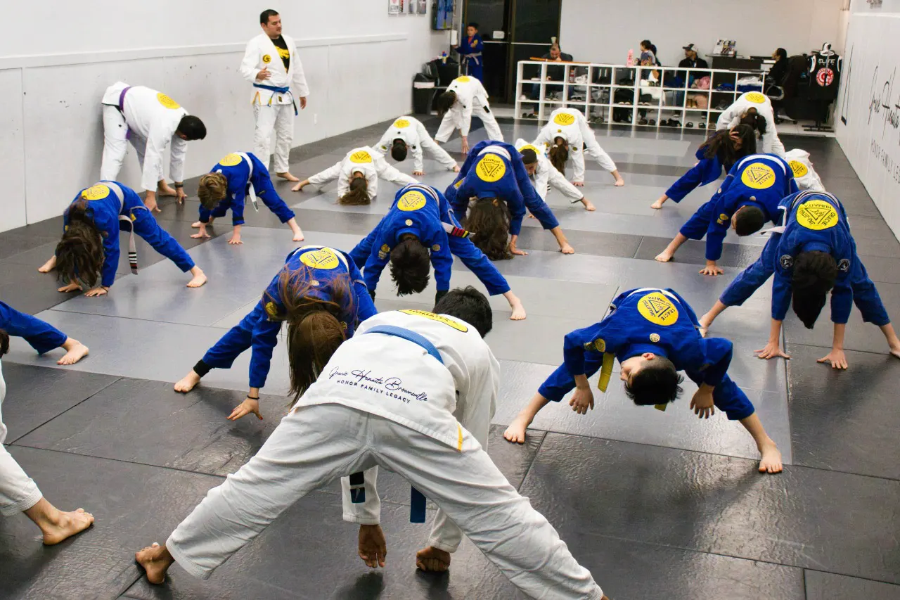 Around twenty children in blue and white jiu-jitsu gis stretching in a wide-legged warm-up while an instructor watches from the front of the mat