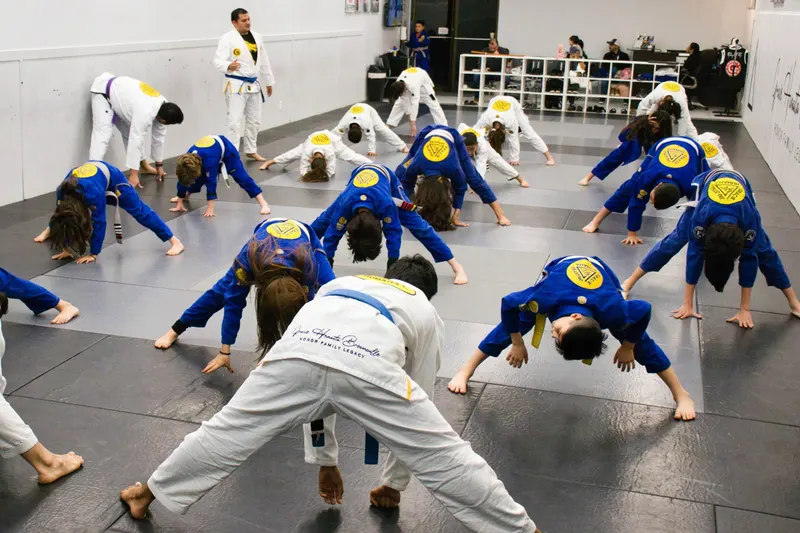 Around twenty children in blue and white jiu-jitsu gis stretching in a wide-legged warm-up while an instructor watches from the front of the mat
