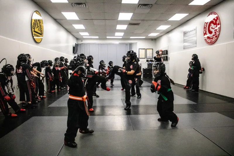 A large Tae Kwon Do class in full sparring gear — padded helmets, gloves and foot guards — training across the mat between the Gracie Humaita and Galvan’s Martial Arts wall crests